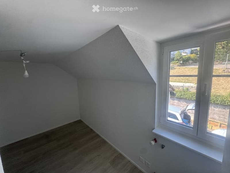 Empty attic room with wooden flooring, white walls, and a large window offering a view of the outside. The room features a sloped ceiling and a single light fixture.