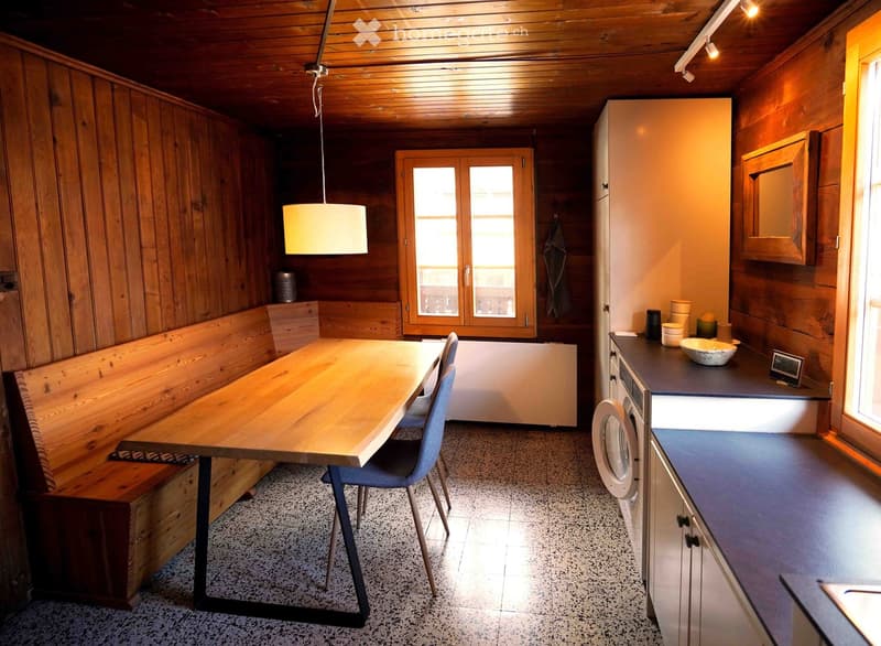 A cozy, wooden-paneled room with a dining area and a small kitchen. Features a wooden table with chairs, a bench seating, a washing machine, and a kitchen counter with various items.