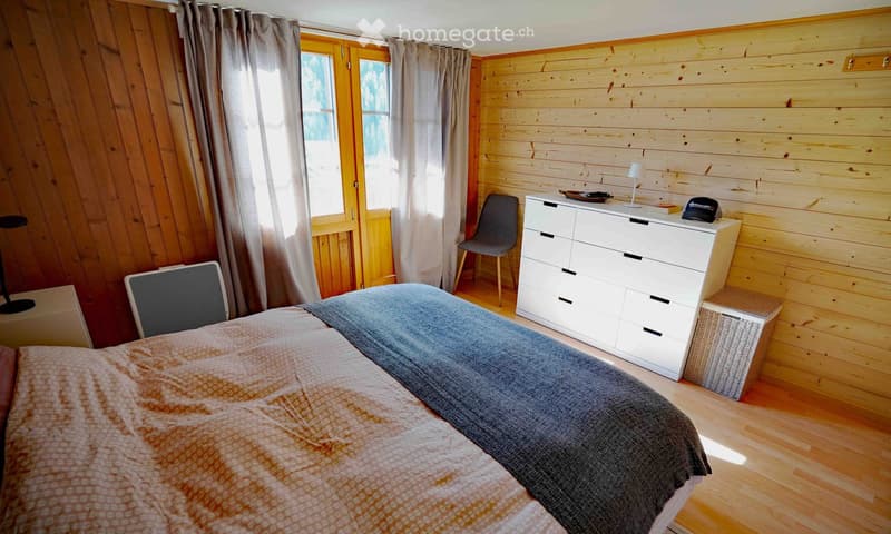 Cozy bedroom with wooden walls and floor, featuring a bed with patterned bedding, a white dresser with drawers, a chair, and a small lamp on the dresser.