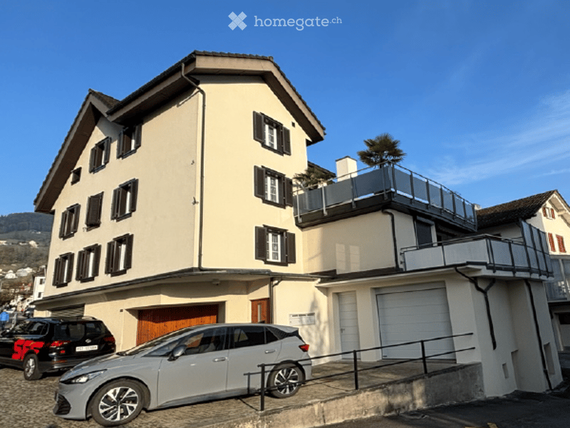 Two-story residential building with beige facade, multiple windows, garage, and balcony on the upper floor.