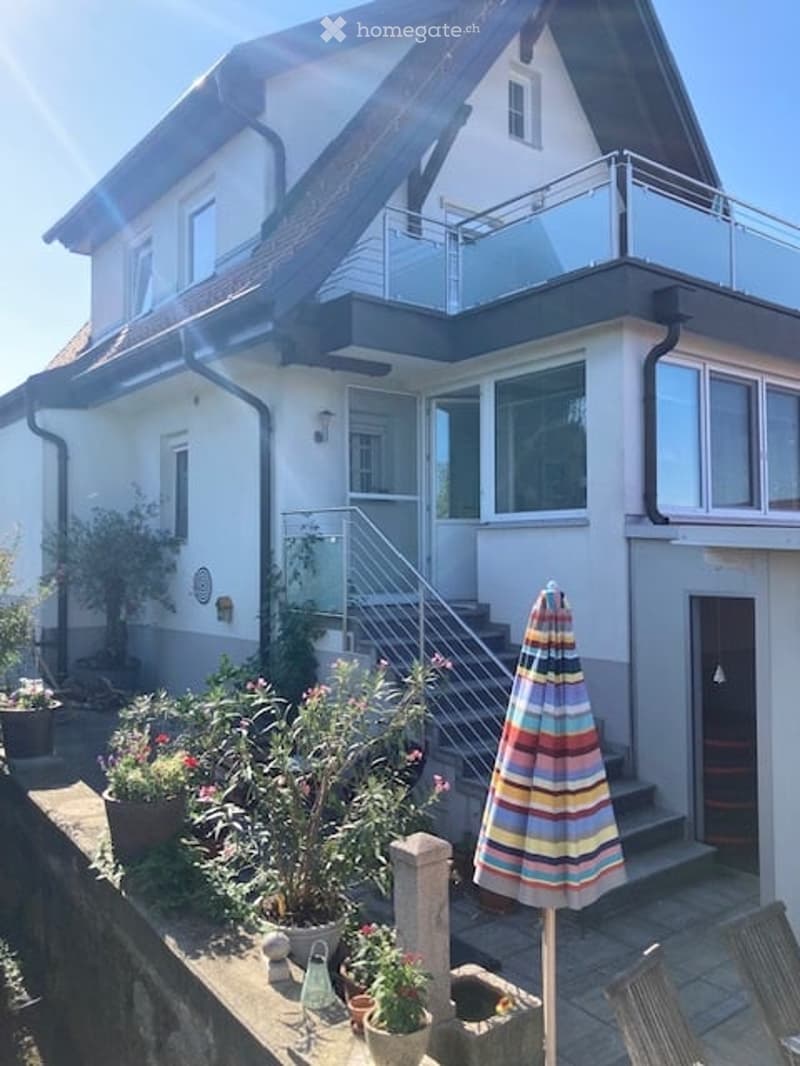 Two-story house with a balcony, large windows, and a well-maintained garden area with potted plants and an umbrella.