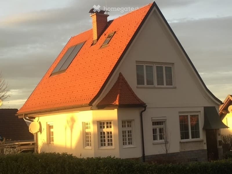 Two-story house with a red-tiled roof, multiple windows, and a chimney. The house has a light-colored exterior and is surrounded by a hedge.