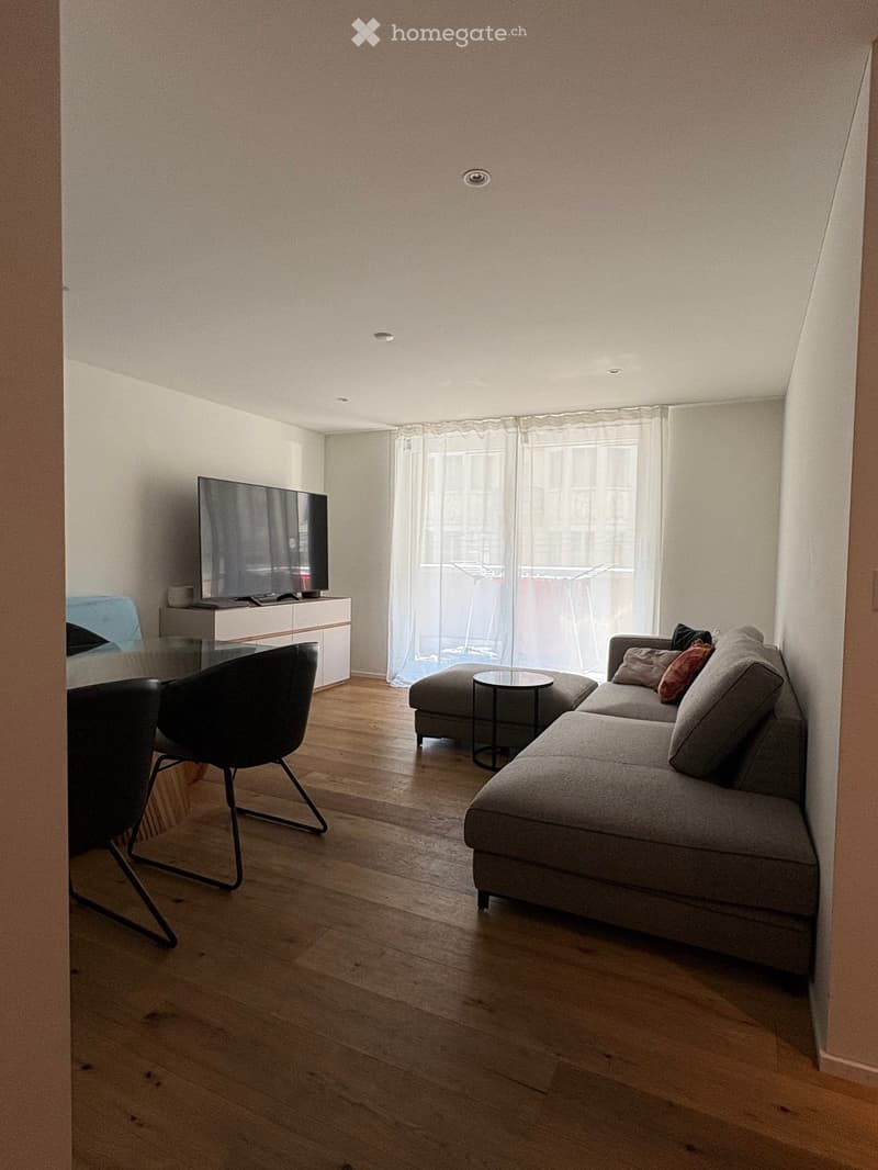 Modern living room with wooden flooring, grey sectional sofa, ottoman, coffee table, and a television on a white cabinet.