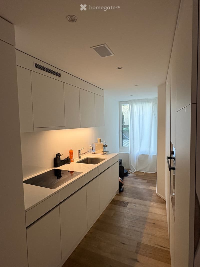 Modern kitchen with white cabinets, wooden flooring, and a small window with white curtains. Features a sink, faucet, coffee maker, and induction cooktop.