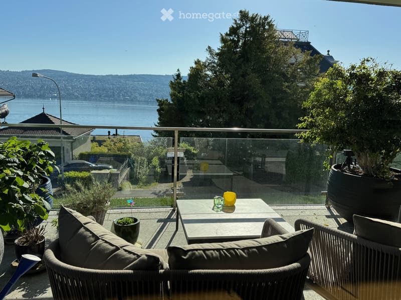 Balcony with a view of the water and mountains, featuring a table, chairs, and potted plants.