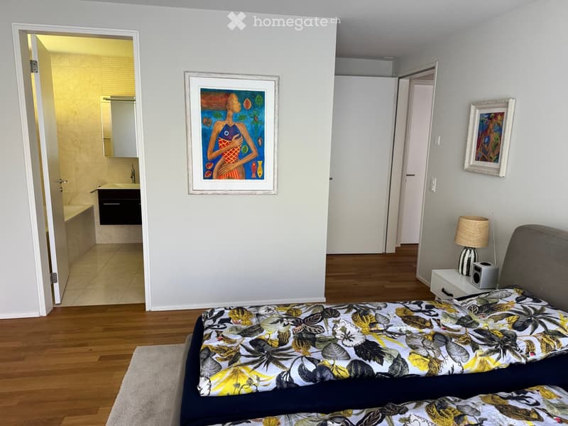 Modern bedroom with wooden flooring, a large bed with a floral duvet, a nightstand with a lamp, and two picture frames on the walls. An open doorway leads to a bathroom with a bathtub and sink.