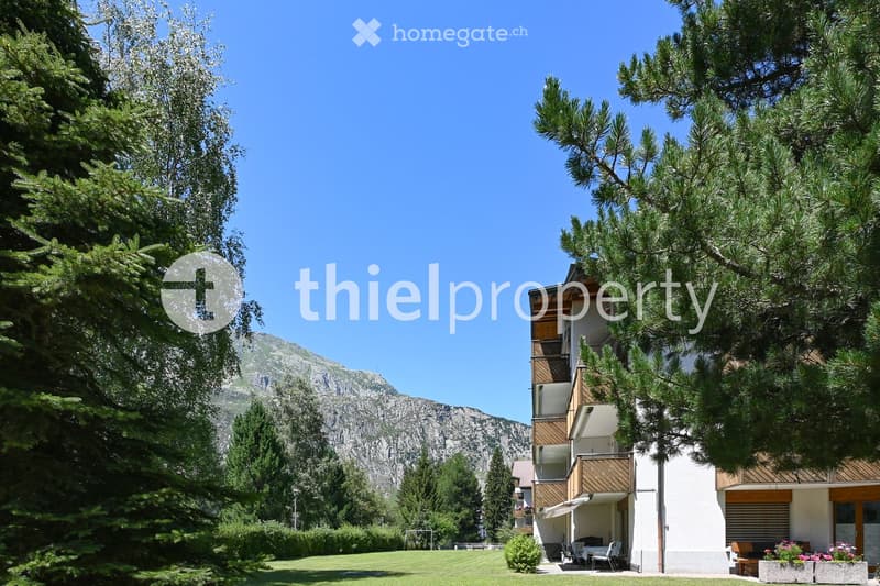 A modern two-story building with balconies, surrounded by lush greenery and a clear view of mountains in the background.