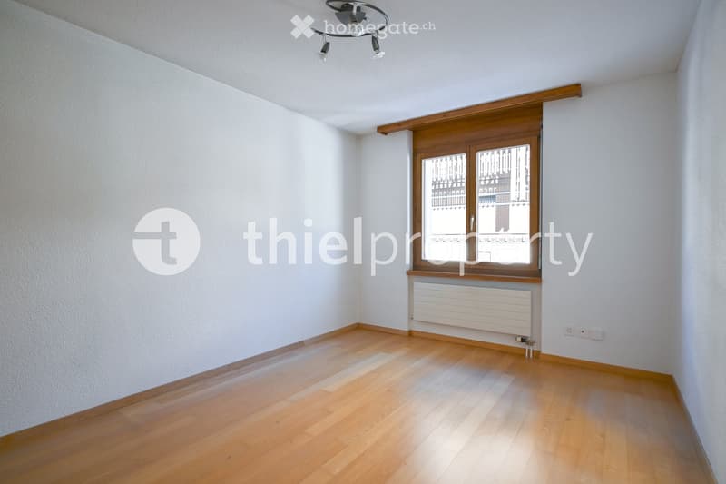 Empty room with wooden flooring, white walls, a window with wooden frame, radiator, and a ceiling light fixture.