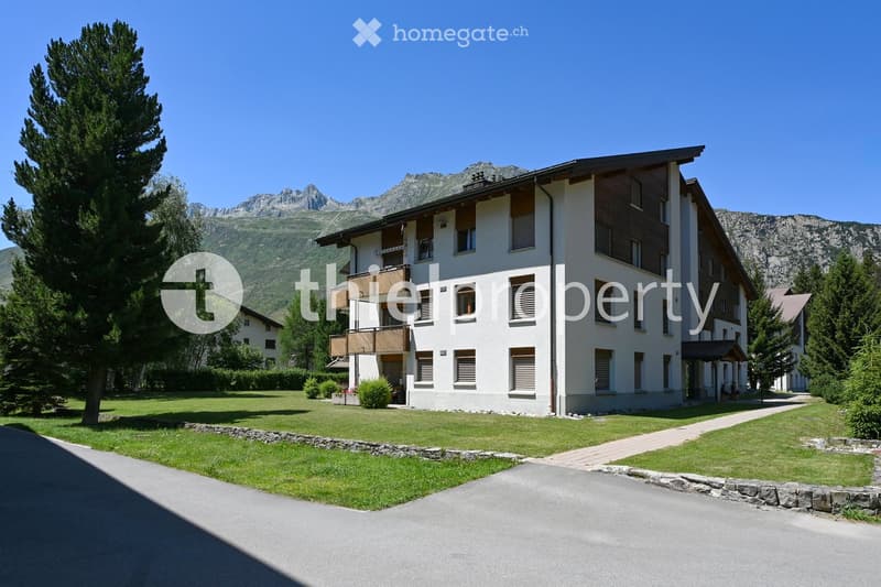 A modern multi-story building with white walls and wooden accents, surrounded by a well-maintained lawn and trees, with a scenic mountain backdrop.