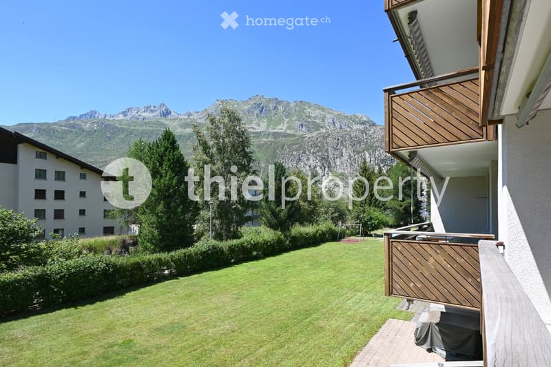 Well-maintained garden with green lawn, trimmed hedges, and trees, with a wooden balcony and scenic mountain view in the background.