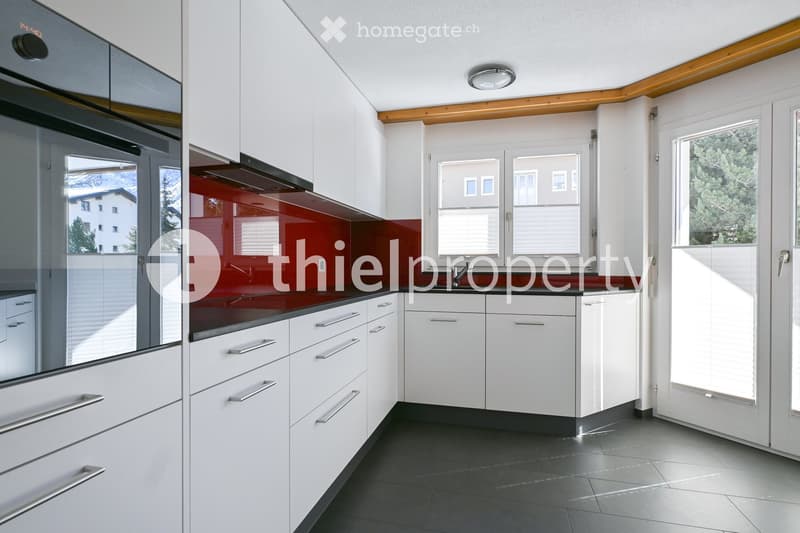 Modern kitchen with white cabinets, stainless steel handles, dark countertops, and a red backsplash. Features a large window and a built-in oven with a glass door.