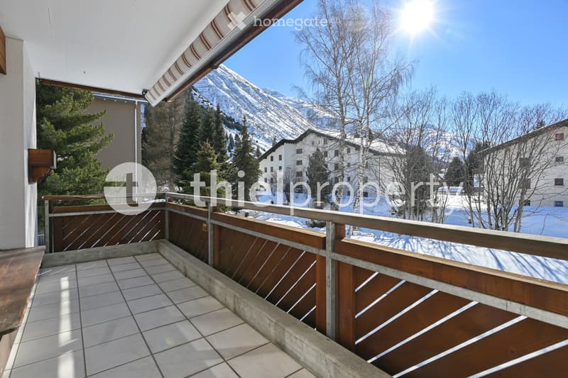 Balcony with wooden railing, tiled floor, overlooking snow-capped mountains and a village.