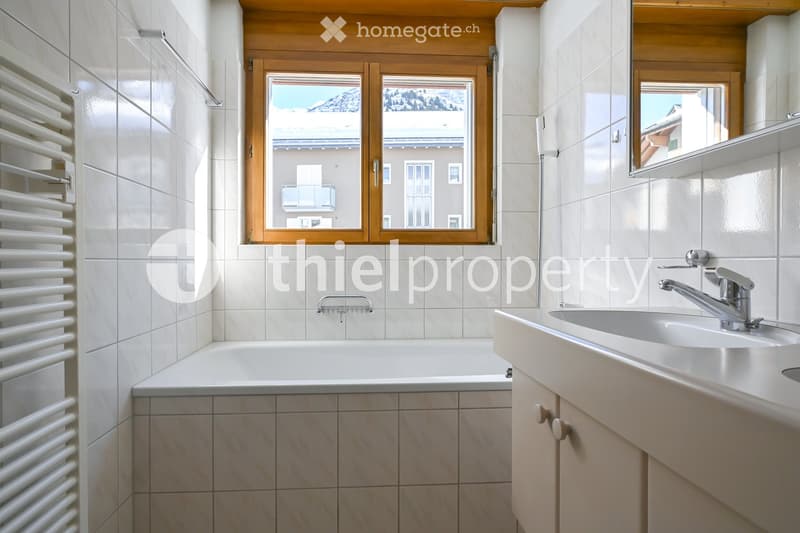 Modern bathroom with a bathtub, sink, and mirror, featuring white tiles and a wooden window frame with a scenic view.