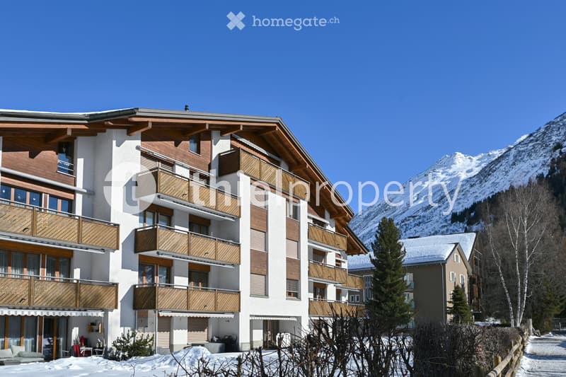 A multi-story residential building with wooden balconies and white walls, situated in a snowy mountain landscape with snow-covered trees and distant peaks.