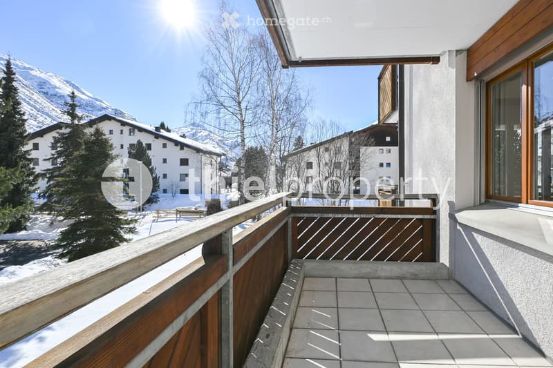 Balcony with wooden railing and tiled floor, overlooking a snowy mountain landscape and neighboring buildings.