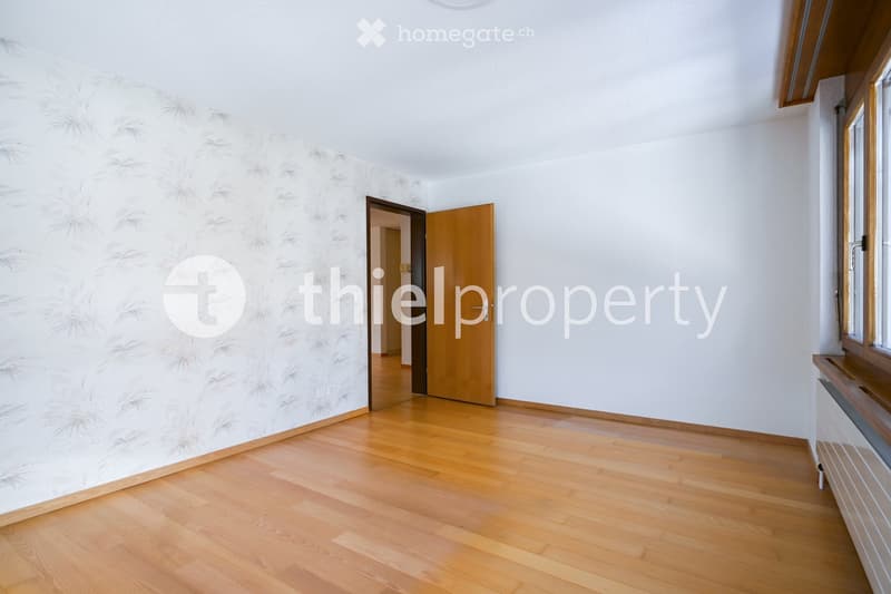 Empty room with light wooden flooring, white walls, and a large window with wooden frame. A door leads to another room.