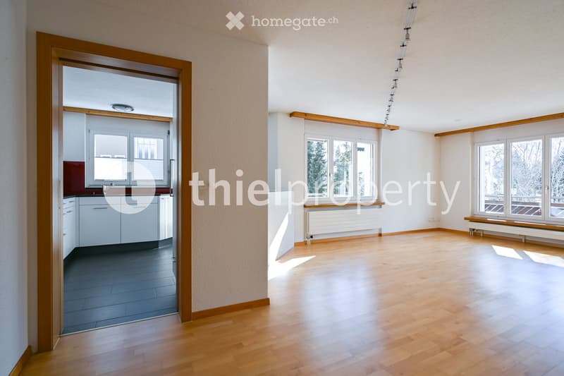 Empty room with wooden flooring, white walls, large windows, and a doorway leading to a kitchen area.