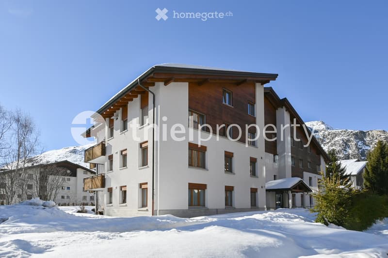 A multi-story residential building with white and wooden facades, multiple windows, and balconies, surrounded by snow-covered ground and mountains in the background.