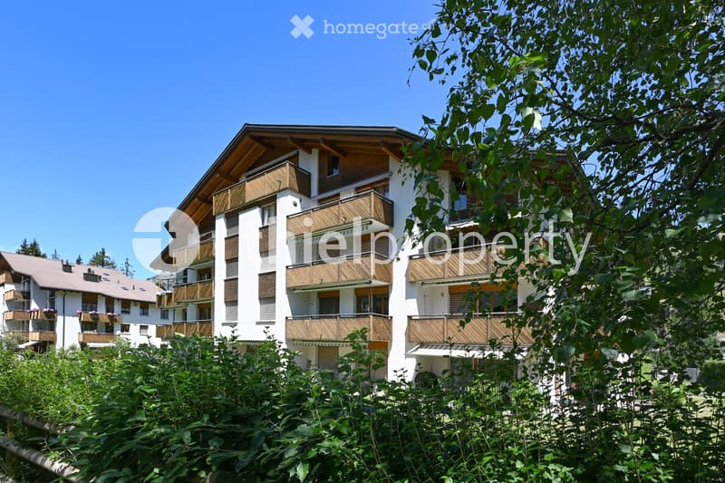 A multi-story residential building with white walls, wooden balconies, and a sloped roof, surrounded by greenery.