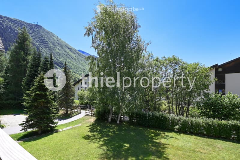 A well-maintained garden with a green lawn, surrounded by trees and shrubs, with a pathway leading to a wooden fence and a view of a mountain in the background.