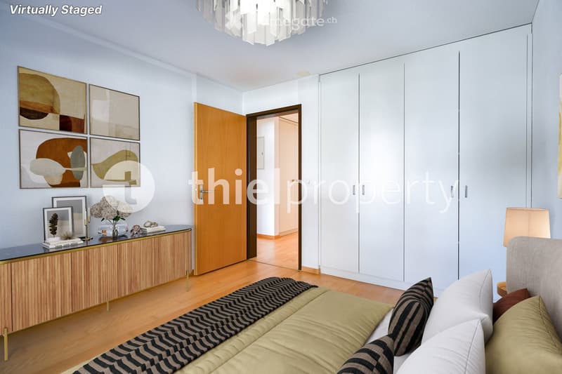 Modern bedroom with a large bed, decorative pillows, a striped blanket, a wooden cabinet with a mirror, and a vase with flowers on the cabinet. The room features light-colored walls, wooden flooring, and a large built-in wardrobe.