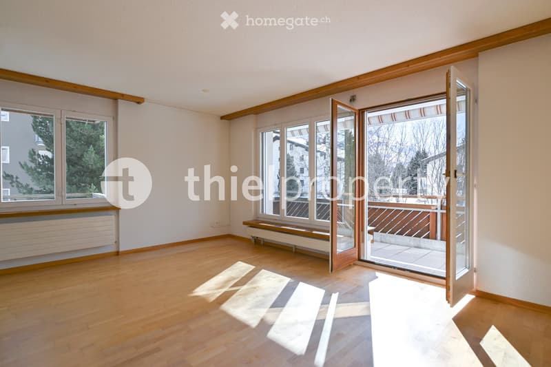 Empty room with wooden flooring, large windows, and a sliding glass door leading to a balcony. The walls are painted white, and the room has a radiator and a bench near the windows.