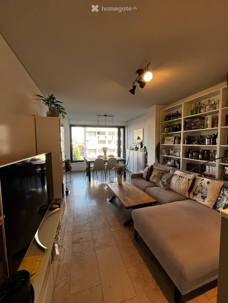 A modern living room with a grey sectional sofa, wooden coffee table, dining area with chairs and table, shelves with books and decorative items, and large windows offering a view of the city.