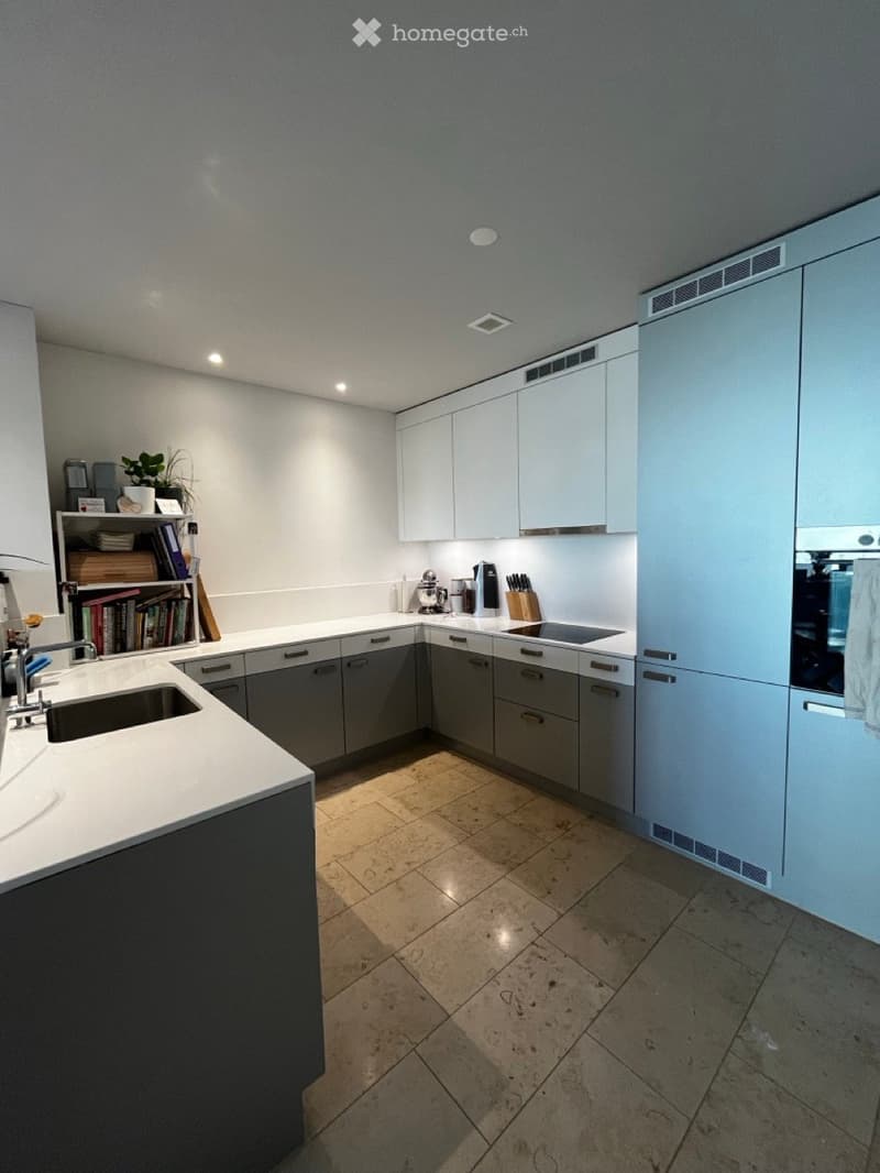Modern kitchen with white and grey cabinets, a white countertop, and a tiled floor. Features include a sink, faucet, oven, bookshelf with books and a plant, blender, coffee maker, and knife block.
