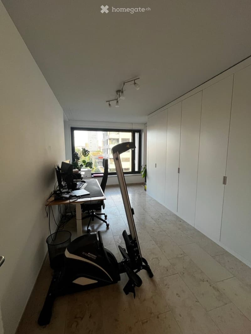 A modern, well-lit room with a desk setup, exercise bike, and large window offering a city view.
