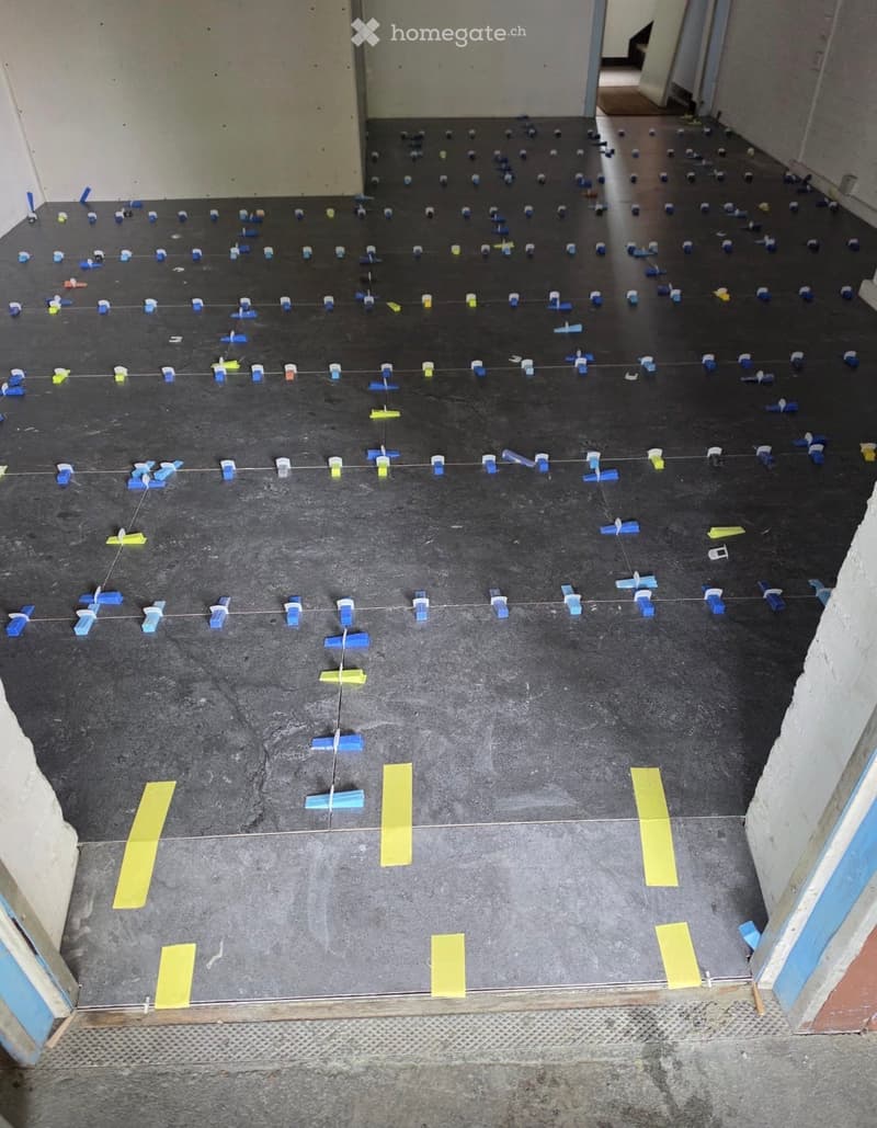 Empty room with a dark grey floor, marked with blue and yellow spacers for tile installation. No furniture or equipment present.