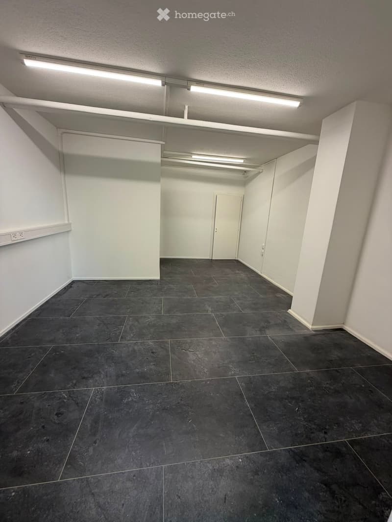 Empty room with dark tiled floor and white walls, featuring a single door and fluorescent lighting.
