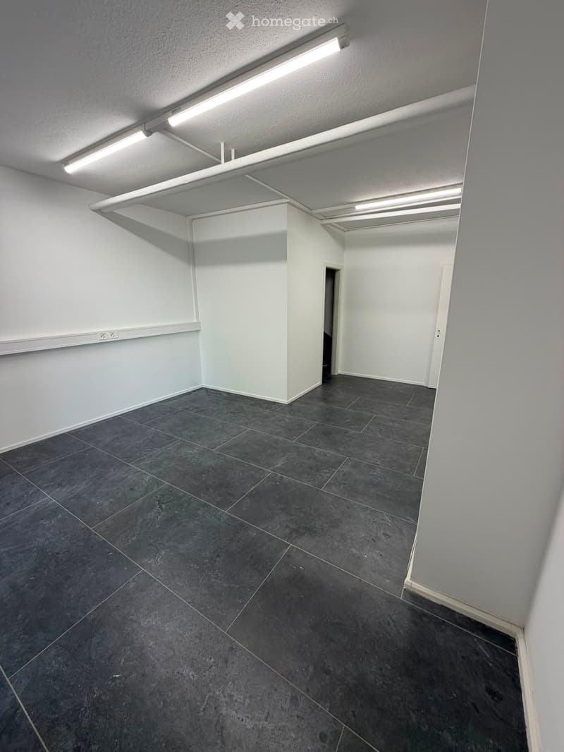 Empty room with dark grey tiled floor and white walls, featuring a single door and fluorescent lighting.