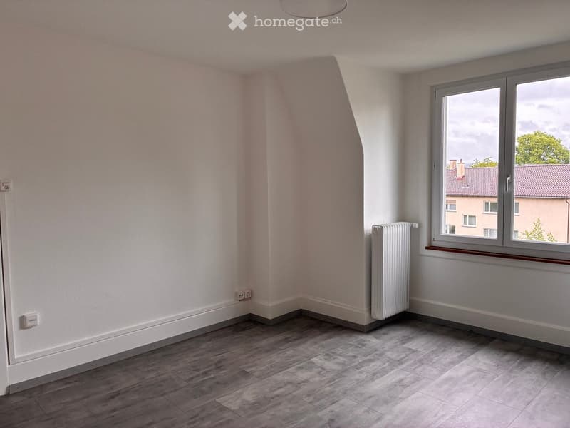 Empty room with light-colored walls, wooden flooring, and a radiator near the window. The window offers a view of neighboring buildings and trees.