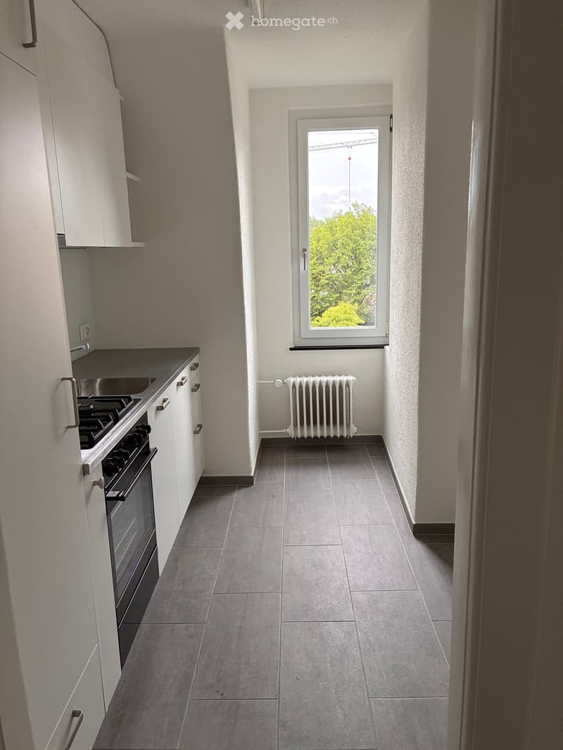 Modern kitchen with white cabinets, dark grey tiles, a stove, sink, and a radiator under a window with a view of greenery.