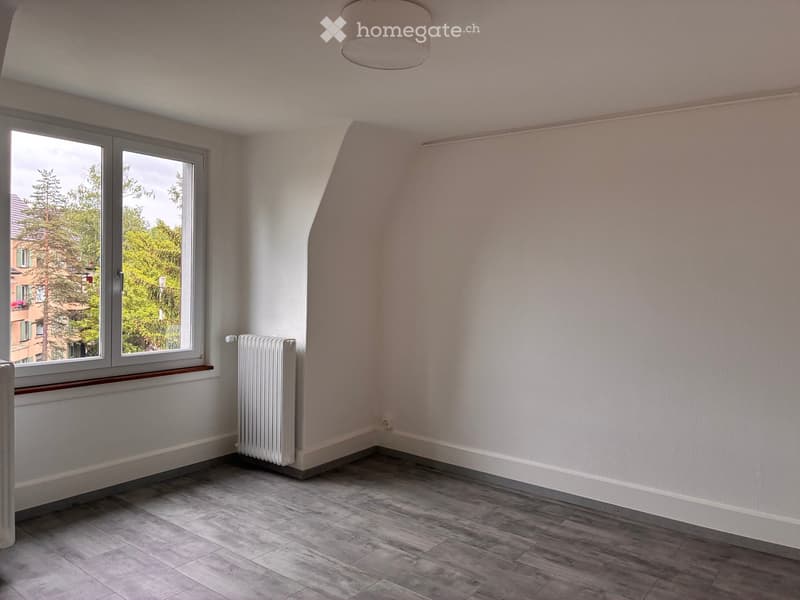 Empty room with light-colored walls, wooden flooring, and a large window with a view of trees and neighboring buildings.