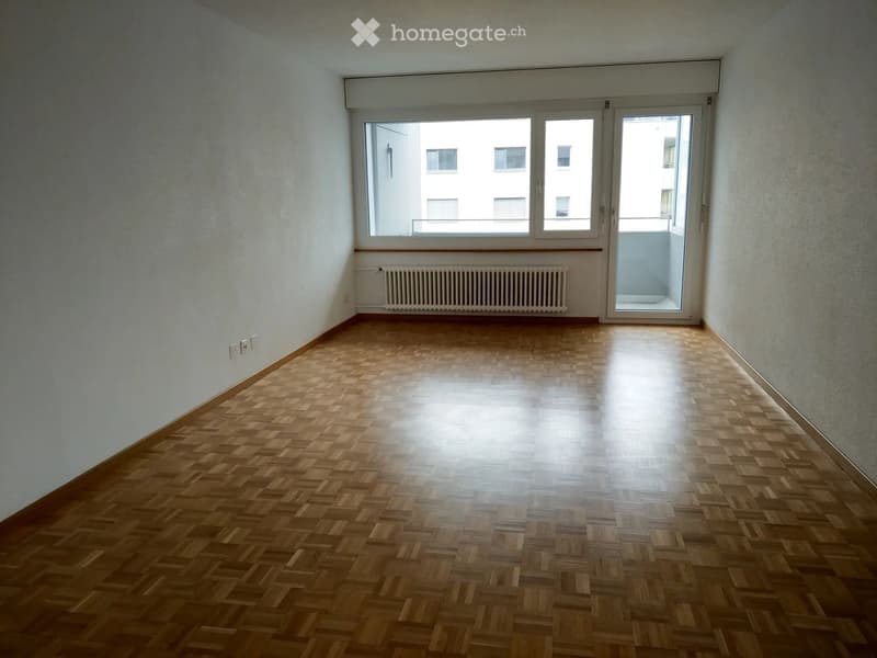 Empty room with wooden parquet flooring, white walls, large window with view, radiator, and sliding glass door leading to a balcony.