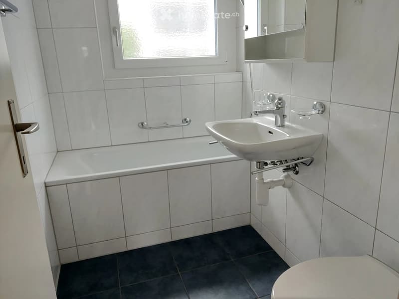 Modern bathroom with white tiled walls, a bathtub, a sink with a modern faucet, a mirror, and a toilet. The room features a window with frosted glass and a door with a silver handle.