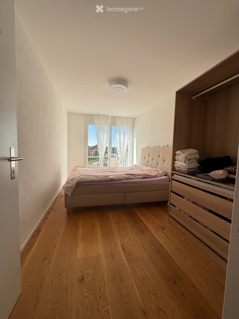Bedroom with wooden flooring, a bed with a tufted headboard, a wardrobe with drawers, and a window with white curtains.