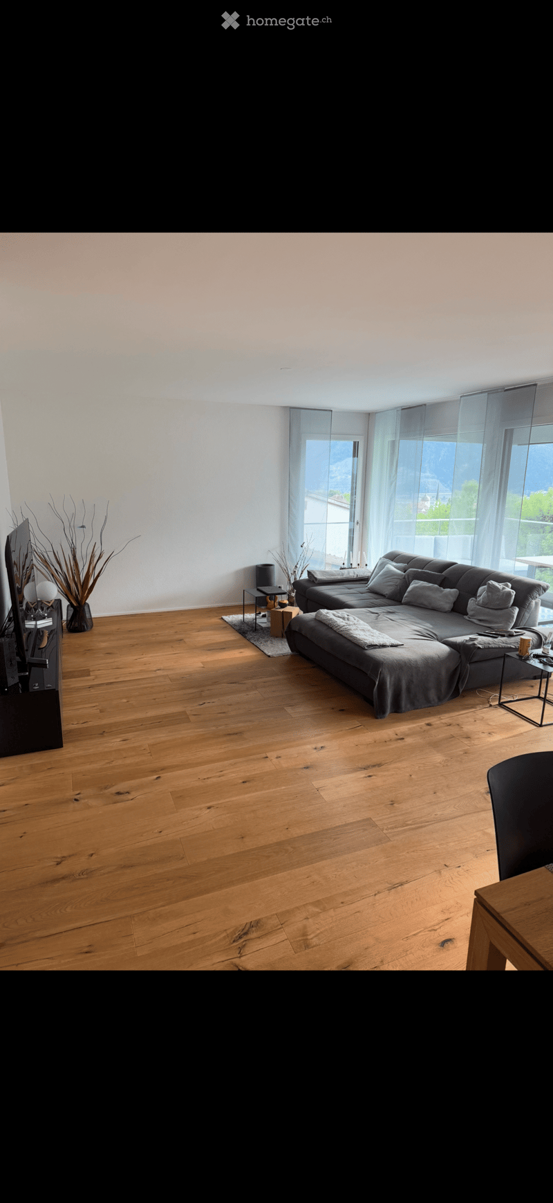 Modern living room with wooden flooring, large windows, grey sectional sofa, coffee table, side tables, and a television stand.