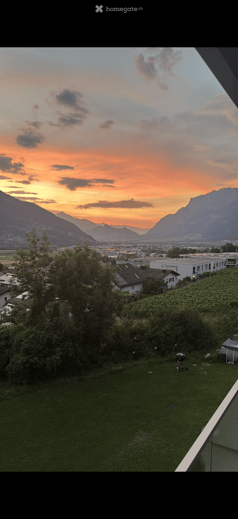 Garden with green grass, trees, and a view of mountains and cityscape at sunset.