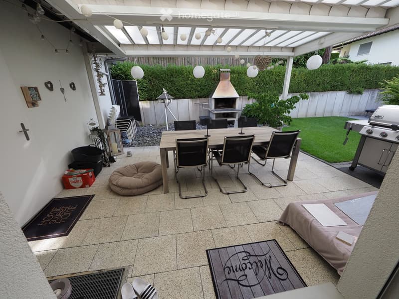 Well-maintained outdoor patio with a dining table, chairs, dog bed, rugs, grill, and fireplace, surrounded by greenery and a covered pergola.