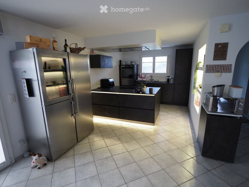 Modern kitchen with stainless steel refrigerator, dark wood cabinets, tiled floor, and various appliances including oven, sink, toaster, and microwave. A small toy is placed on the floor near the window.