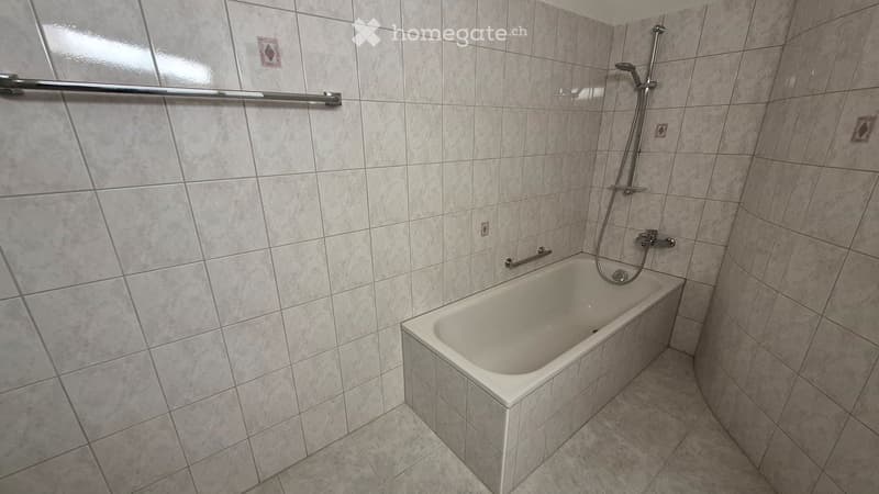 Bathroom with a white bathtub, tiled walls and floor, and a showerhead with a rail.