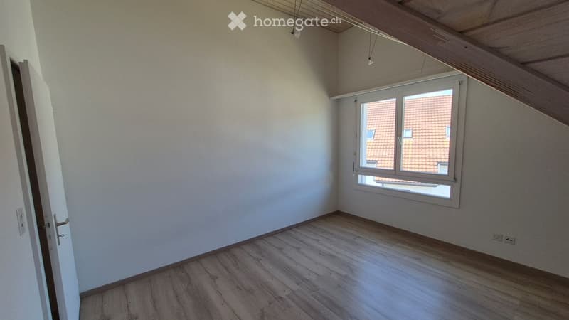 Empty room with wooden flooring, white walls, and a window showing a neighboring building with a red-tiled roof.