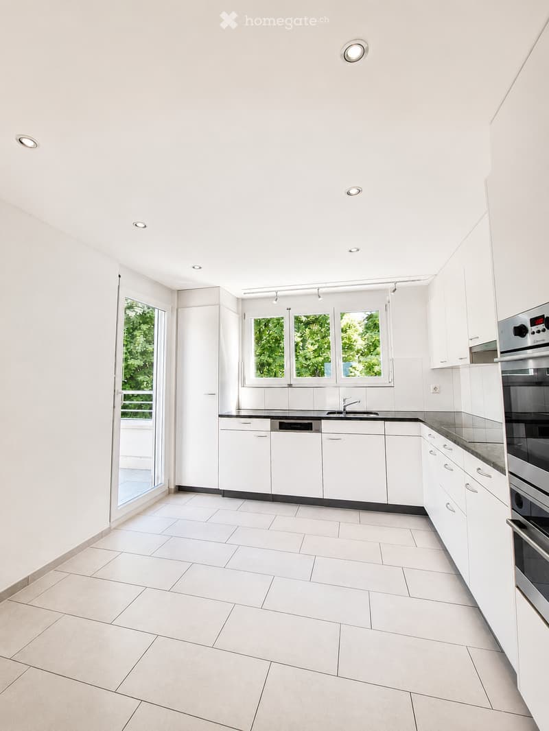 Modern, minimalist kitchen with white cabinets, tiled floor, and stainless steel appliances including an oven and dishwasher. Features large windows and recessed lighting.