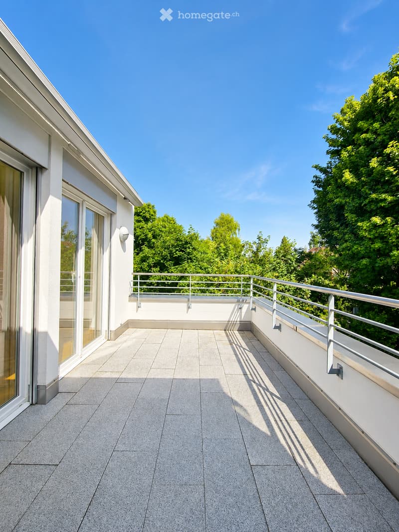 Spacious balcony with modern tiled flooring, white railing, and large windows overlooking a green, tree-filled area.