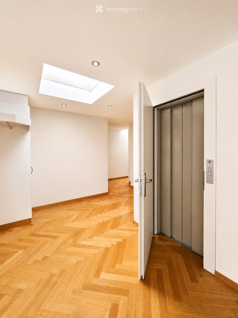 Bright, minimalist hallway with herringbone wooden flooring, white walls, recessed lighting, and a skylight above. Features an elevator on the right side.