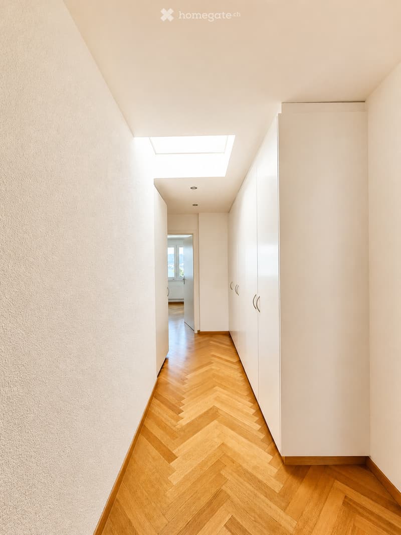 A modern hallway with herringbone wooden flooring, white walls, built-in wardrobes, and a skylight at the top.