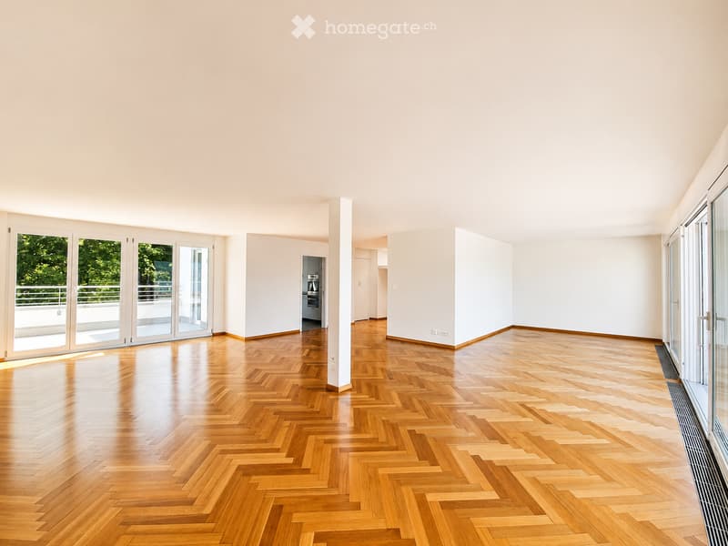 Empty room with light-colored walls and polished wooden herringbone flooring. Large glass sliding doors lead to a balcony with a metal railing and greenery visible outside.