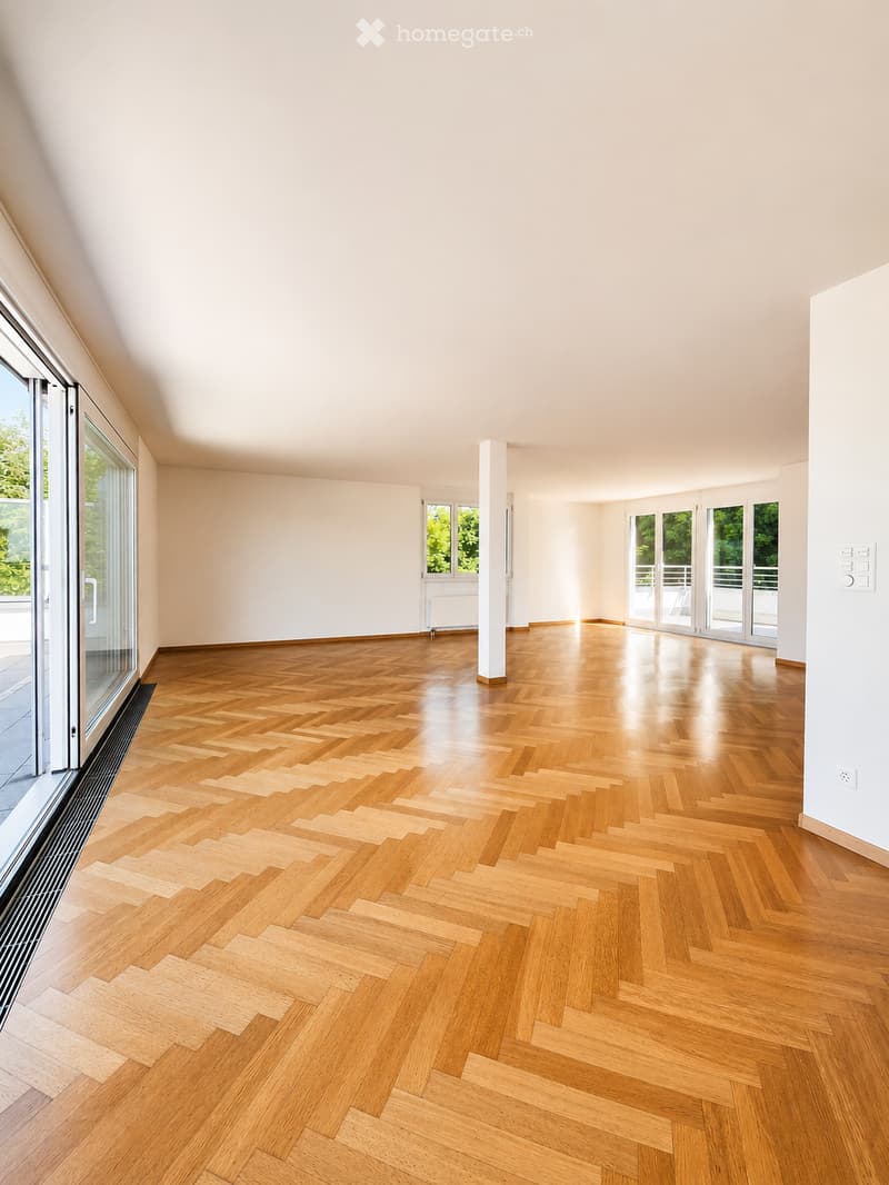 Empty room with light wooden herringbone flooring, white walls, large windows, and a central column.